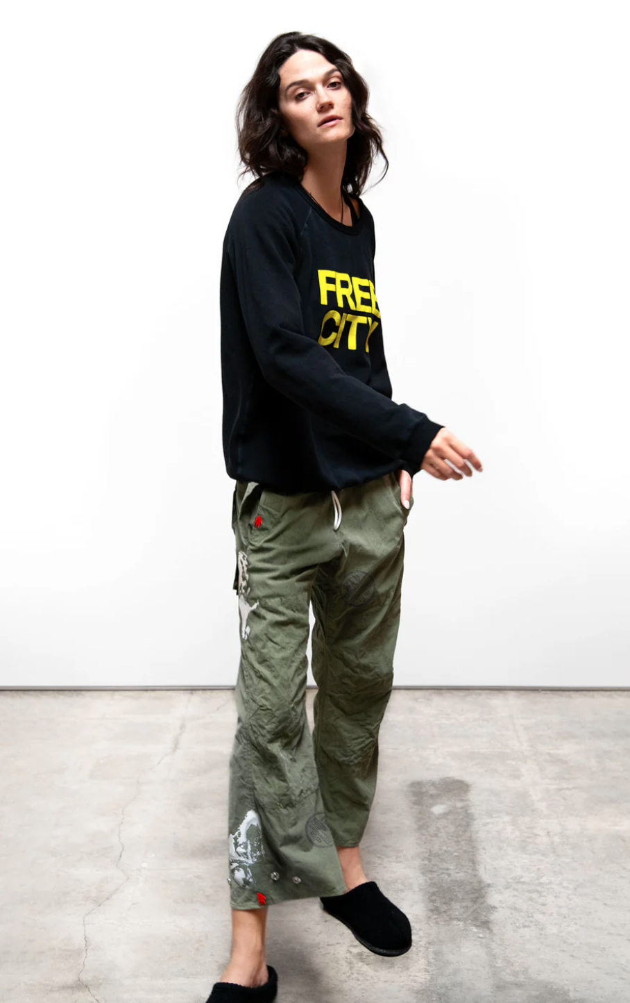 A woman in a casual black hoodie made of French terry and patterned green cargo pants stands against a plain backdrop, looking to the side with a relaxed pose wearing the FREECITY SUPERYUMM BIGGY RAGLAN from Free City (sparrow, LLC).