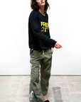 A woman in a casual black hoodie made of French terry and patterned green cargo pants stands against a plain backdrop, looking to the side with a relaxed pose wearing the FREECITY SUPERYUMM BIGGY RAGLAN from Free City (sparrow, LLC).