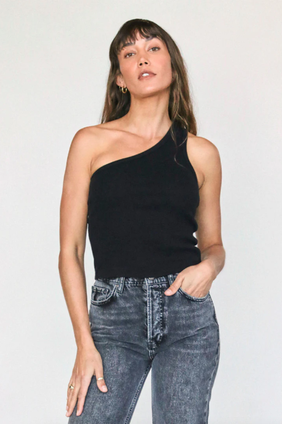 A woman wearing a Perfectwhitetee Call Me One Shoulder Structured Rib Tank and blue jeans stands confidently against a plain white background in her bungalow. Her hand rests on her hip, and she has a relaxed expression with bangs framing her face.