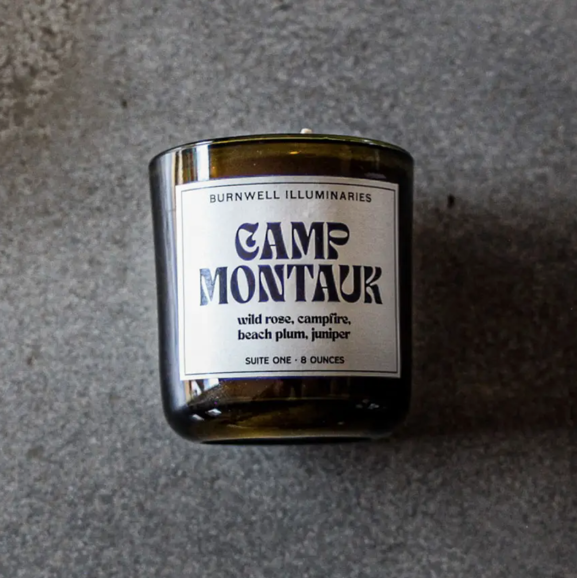 A Barnaby Black Candle in a dark glass jar labeled &quot;Camp Montauk&quot; with scent notes of wild rose, campfire, beach plum, and juniper listed, placed on a gray textured surface in Arizona style. (Brand: Faire)