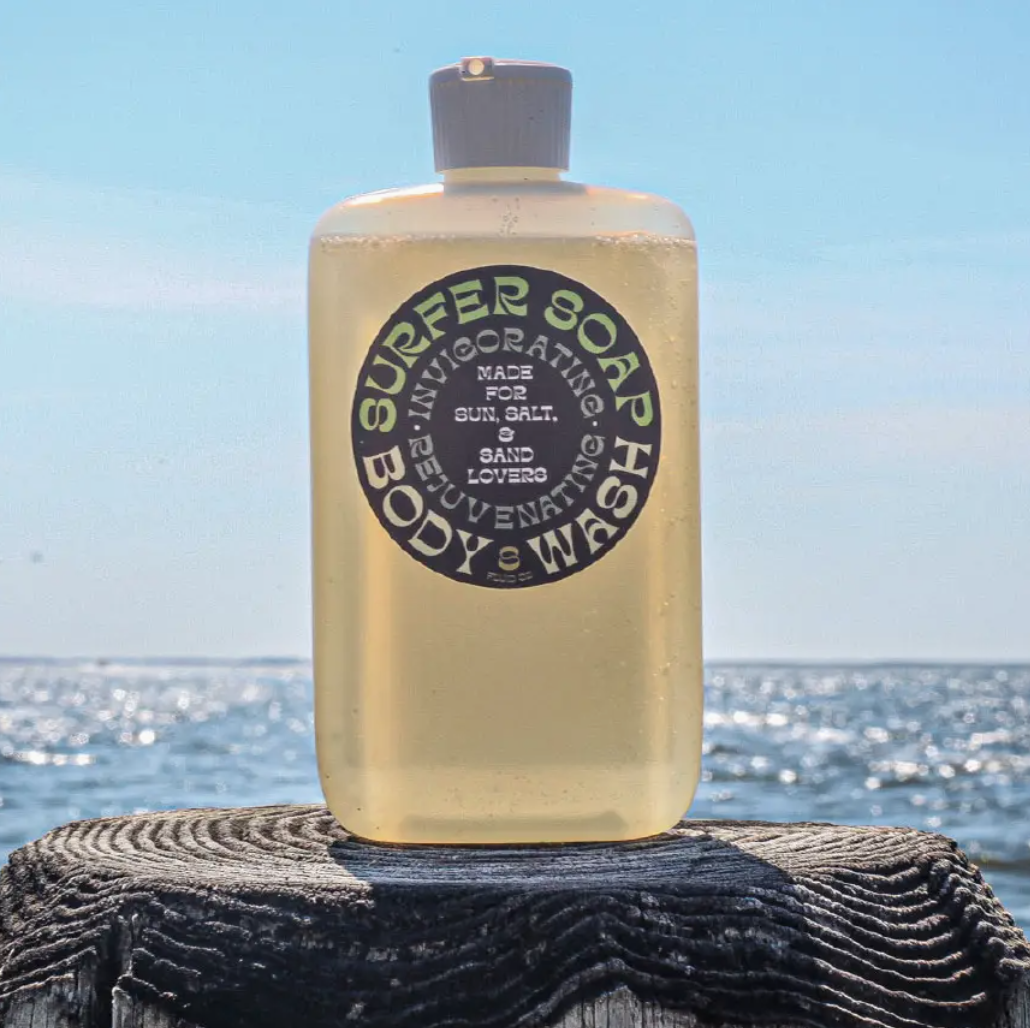 A translucent bottle of Faire body wash resting on a weathered wooden beam against a backdrop of a sunlit ocean. The label indicates it is vegan and biodegradable. Its style echoes the casual, beachy vibe of an Arizona bungalow.