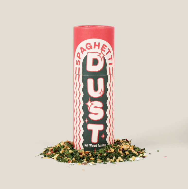 A tall cylindrical container labeled &quot;Faire Spaghetti Dust&quot; in bold red and white text, surrounded by a small pile of green and yellow seasoning at the base, styled like a classic Arizona bungalow, against a plain light background.