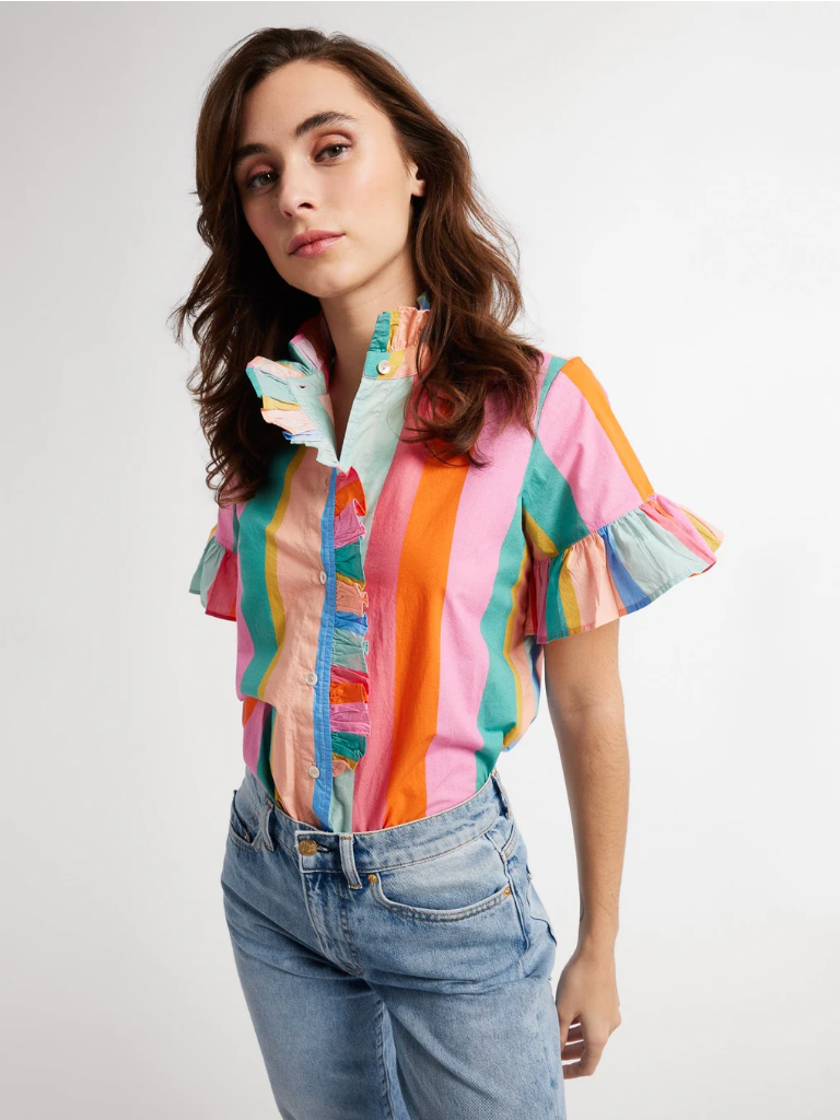 A woman with long brown hair wearing a Mille Vanessa Top, a colorful striped blouse with cascading ruffles, and blue jeans, posing against a light background.