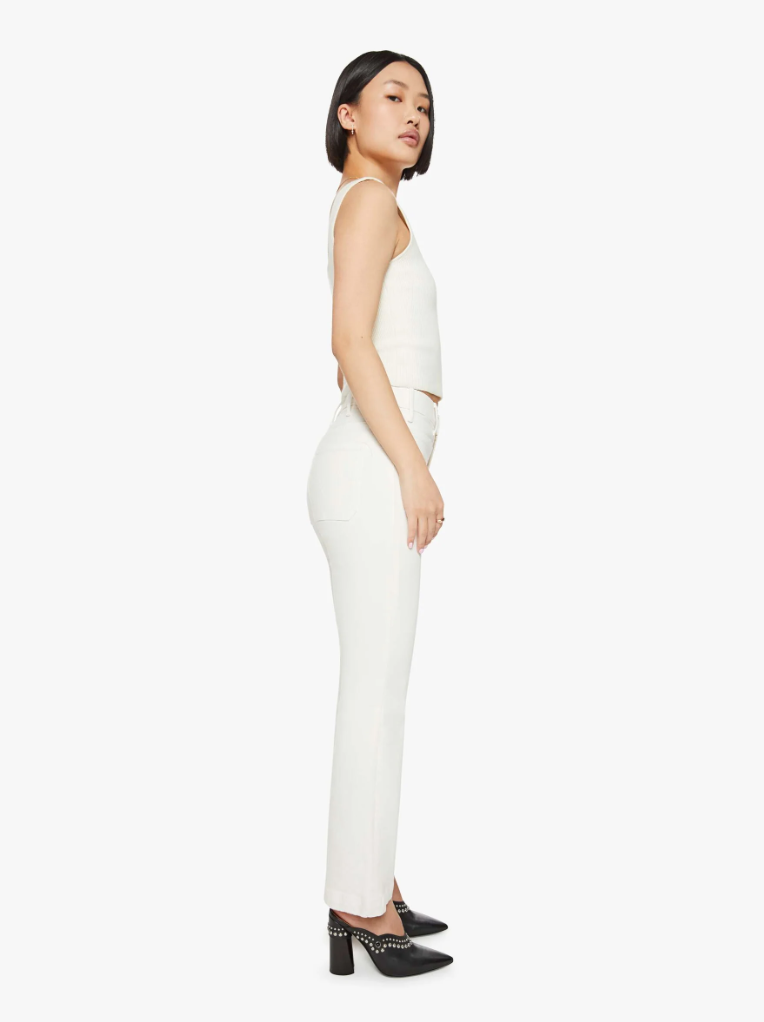 A woman wearing Mother's The Hustler Patch Pocket Flood white sleeveless top and white high-rise bootcut pants, standing sideways, looking over her shoulder. She has short black hair and is wearing black heels.