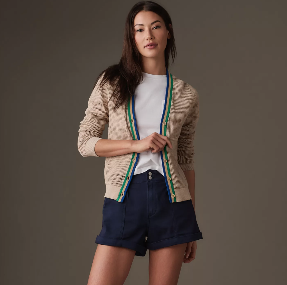 A woman stands confidently in a Scottsdale studio, wearing a Kule Zoey Cardigan with green and blue trim, a white top, and navy shorts. Her posture is relaxed and her expression is serene.