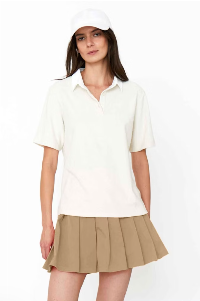 A young woman wearing a Kule cotton terry polo shirt and a khaki pleated skirt with a white cap, standing against a plain white background. She appears confident and casually stylish.