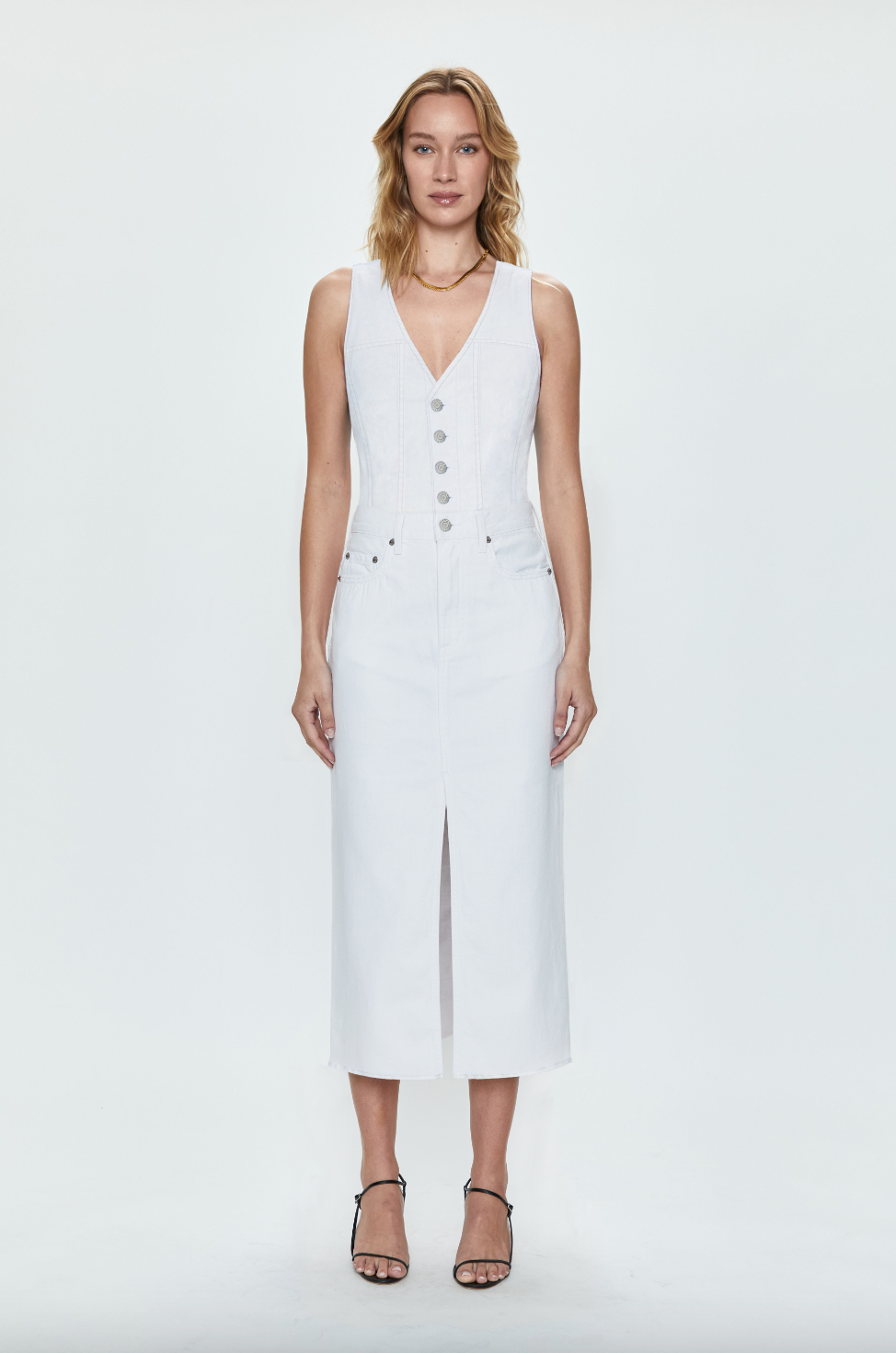 A Los Angeles-based woman wearing a stylish white sleeveless Alex Dress by Pistola with buttons stands against a clean white background. She has shoulder-length blonde hair and is wearing black strappy sandals.