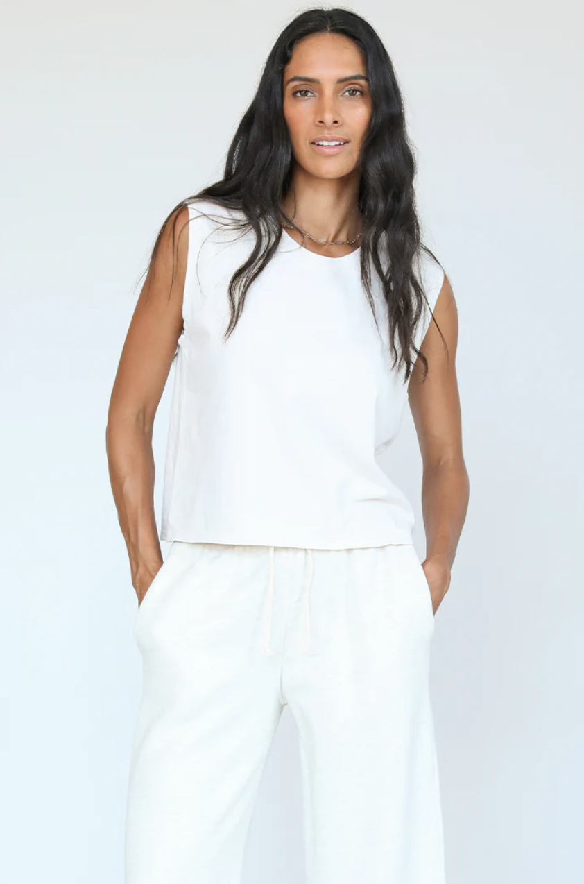 A woman wearing a white Perfectwhitetee Miley cotton jersey muscle tank and matching pants stands against a light gray background in her bungalow, looking directly at the camera with a neutral expression.