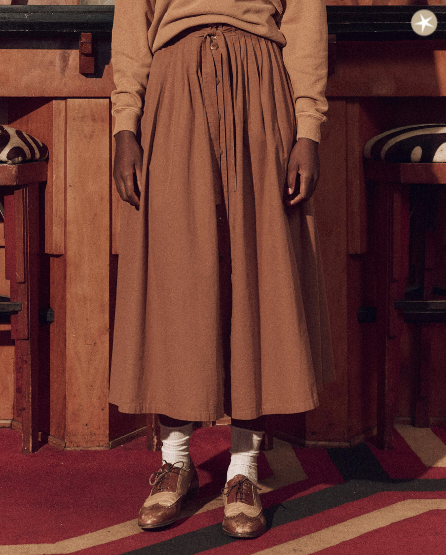 A person stands wearing The Treeline Skirt Suntan paired with a matching sweater, brown and beige brogues, and white socks, shown from the waist down in a rustic bungalow interior setting.