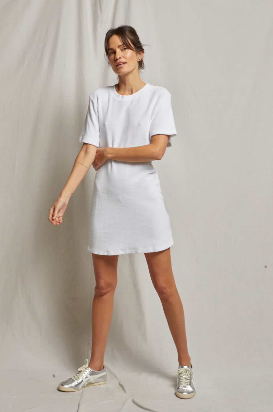A woman in a Perfectwhitetee EMELIA loop terry tee dress and metallic sneakers posing casually against a bungalow's neutral backdrop, looking effortlessly chic.