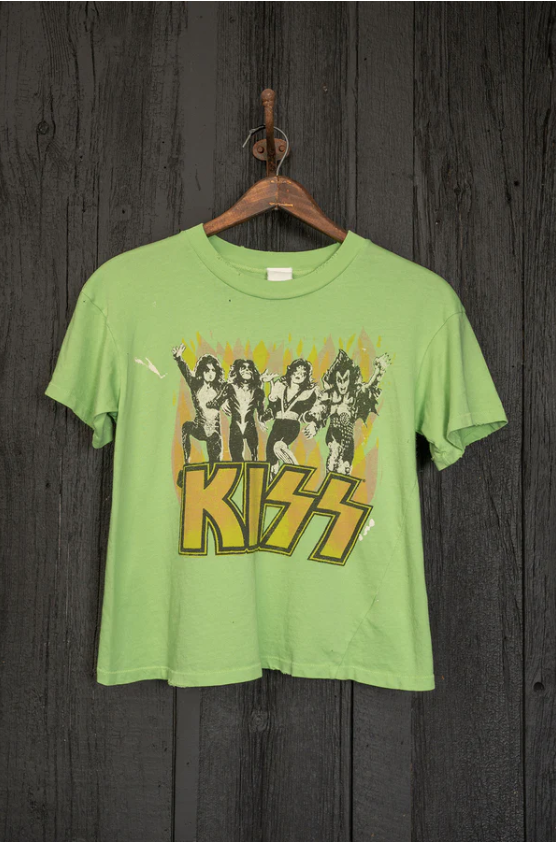 A green Made Worn Kiss Crop Tee Electric with the band KISS graphic printed on the front, hanging on a rustic metal hook against a dark wooden backdrop in Arizona style.