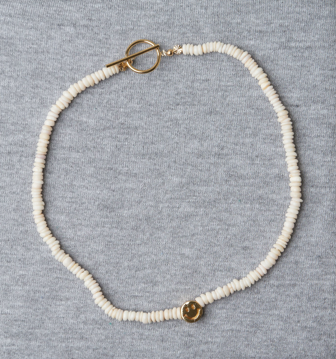An elegant Puka Gold Smiley Choker necklace with white beads and a small gold-filled clasp, displayed on a light gray fabric background by Aquarius Cocktail.