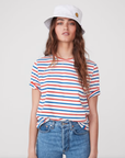 A woman with long brown hair is wearing a white bucket hat with a small smiley face patch, The Modern from Kule featuring red, blue, and white horizontal stripes, and blue jeans. She is standing against a plain white background, looking directly at the camera.