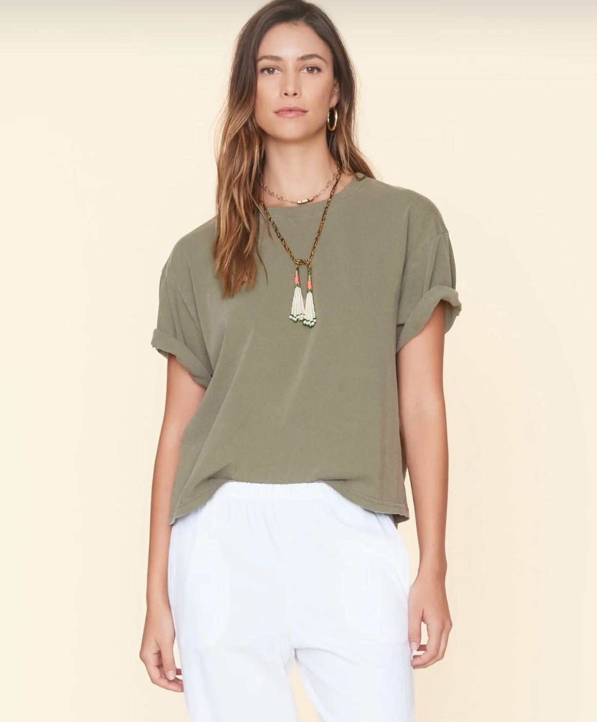 A person with long, brown hair wears a loose-fitting olive green Palmer Tee by Xirena with rolled sleeves, a long beaded necklace with white and green tassels, and white pants. The ultra-soft cotton jersey shirt accentuates their relaxed posture as they stand against a neutral background, looking straight ahead.