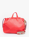 A bright pink drawstring bag with short handles and an adjustable shoulder strap that has "ciao ciao" written on it in red. The strap also features a blue and red design, giving it a lively Arizona style. The background is plain white. This is the Sac Tout from Clare Vivier.