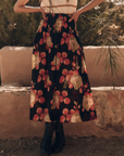 A woman wearing a white top and The Canary Skirt by The Great Inc. poses outside. The skirt, crafted from sundried cotton fabric, features a dark background with large cabbage rose prints in shades of red and yellow. She also wears black lace-up ankle boots. Her upper body is out of the frame.
