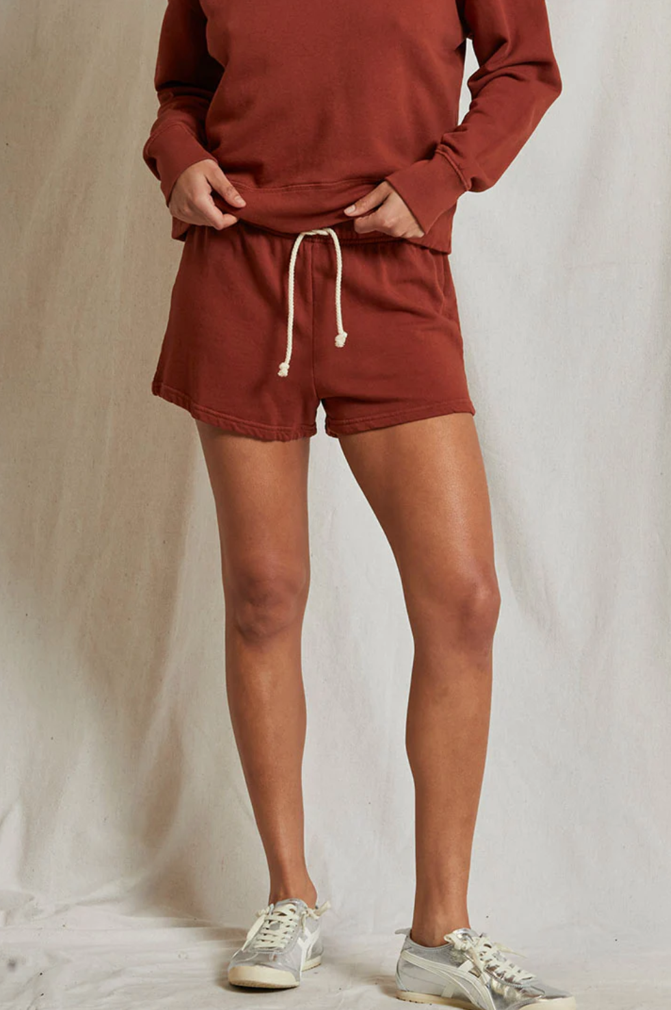 A person stands against a light background wearing the rust-colored Layla Terry Short and a matching sweatshirt, both from Perfectwhitetee. Their hands are on their hips, and they are sporting light-colored sneakers. Made from soft French terry cotton, the outfit exudes cozy comfort. Their face is not visible in the image.