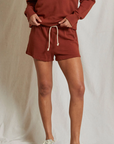 A person stands against a light background wearing the rust-colored Layla Terry Short and a matching sweatshirt, both from Perfectwhitetee. Their hands are on their hips, and they are sporting light-colored sneakers. Made from soft French terry cotton, the outfit exudes cozy comfort. Their face is not visible in the image.