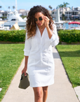 A woman with long curly hair walks on a pathway near a marina, wearing a relaxed fit Southern Dress in Italian Performance Linen by Frank & Eileen with rolled-up sleeves. She holds a small woven handbag in one hand and adjusts her sunglasses with the other. The background includes boats and waterfront homes.