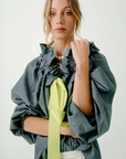 Against a plain background, a light-haired individual stands out in the Beautiful Mess Poet Blouse by Aquarius Cocktail. The grey blouse features grand ruffle details and is adorned with a bright yellow ribbon. Their hair is partially tied back, and they look directly at the camera while holding one of the sleeves with an elastic cuff, subtly revealing a visible tattoo on their forearm.