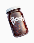 A glass jar with a white lid containing a dark, chunky substance—likely Boon Sauce by Faire. The jar, labeled "BOON" in bold white letters, promises intense flavor and heat. It is positioned at an angle against a plain white background.