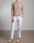 Against a plain backdrop, a person exudes casual style in the Andy Pant by Pharaoh, paired with a light brown short-sleeve top featuring sateen accents. Their ensemble is completed with relaxed-fit white pants and beige slip-on sandals, while one hand rests in their pocket.