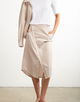 A person in a white t-shirt has tucked it into Pharaoh's Nico Skirt, featuring beige and white vertical stripes, an asymmetric placket, and front slash pockets. Draped over one shoulder is the matching striped shirt. They wear light-colored sandals while standing on a plain surface.
