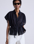 A person in the Agni Floaty Top, a lightweight black button-up blouse with cap sleeves and waist tie, from Apiece Apart, paired with white pants. They have short hair, wear dark sunglasses and elegant drop earrings against a plain gray backdrop.