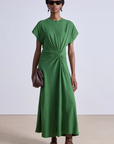 A person wears the Apiece Apart Volta Twist Dress in green organic cotton, featuring short sleeves and a waist twist detail. They accessorize with sunglasses, hoop earrings, and a brown clutch, completing the look with brown strappy sandals.