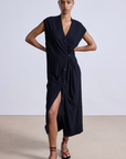 Wearing the Olana Knit Midi by Apiece Apart, someone stands confidently on a beige floor against a plain backdrop. The sleeveless black V-neck dress with buttons and a slit is complemented by brown strappy sandals, creating the perfect lightweight spring layer.