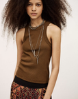 A woman with voluminous curly hair exudes bohemian flair in the sleeveless brown BA&SH Top Cynda, layered silver necklaces, a bracelet, and a geometric patterned skirt, standing against a plain light background.