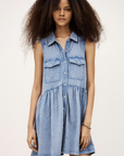 A woman with voluminous, curly hair wears the BA&SH Dress Ilona—a sleeveless, light blue denim mini dress with two chest pockets—facing forward against a plain background.