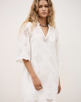 A woman with long wavy blonde hair wears the BA&SH Dress Malo—a loose, knee-length white broderie anglaise dress with lace cutout patterns and elbow-length sleeves—standing against a plain light background.