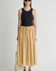 A woman faces forward in sunglasses, a black sleeveless top, and the Apiece Apart Hanne Tie Maxi Skirt— a long yellow A-line style with an elastic waist—paired with black sandals, a headband, and a netted tote against a white background.