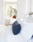 Bright, airy bedroom with white bedding and a navy blue, handwoven POM POM Hendrick Body Pillow in the foreground. Through an open door, another bed with white covers, a rust pillow, and a leafy plant by the window is visible.