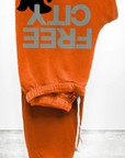 An orange pair of Free City (sparrow, LLC) FREECITY Large Sweatpant made from lightweight French terry, featuring the text "FREE CITY" in large, grey letters and a black silhouette of a small animal on the side. The unisex FLOW sweatpant is folded neatly, showcasing the text and design prominently against a plain grey and white background.