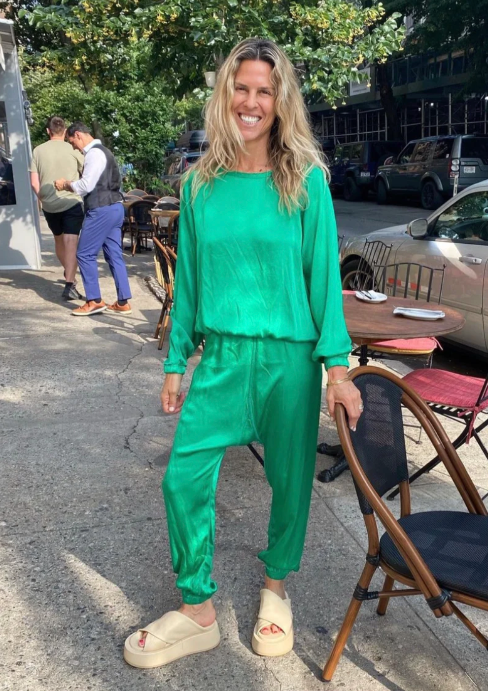 A joyful woman in a vibrant green outfit standing on a sunlit sidewalk in Scottsdale Arizona, with people and outdoor café seating in the background. She wears white shoes and has long, wavy blonde hair while clutching the BOWIE | SATIN Kelly from Aquarius Cocktail.