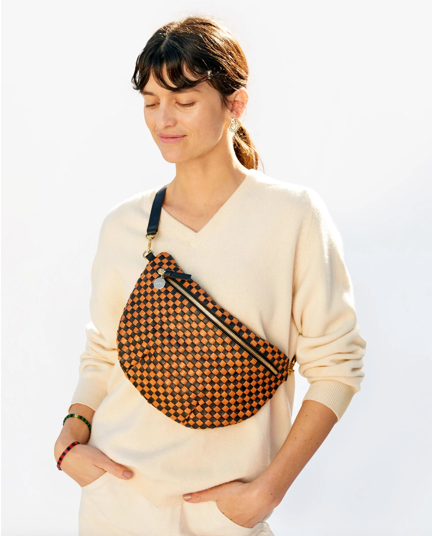 A woman in a white sweater smiles gently with her eyes closed, wearing a Clare Vivier Grande Fanny Woven Checker across her chest against a plain background, capturing the relaxed Arizona style.