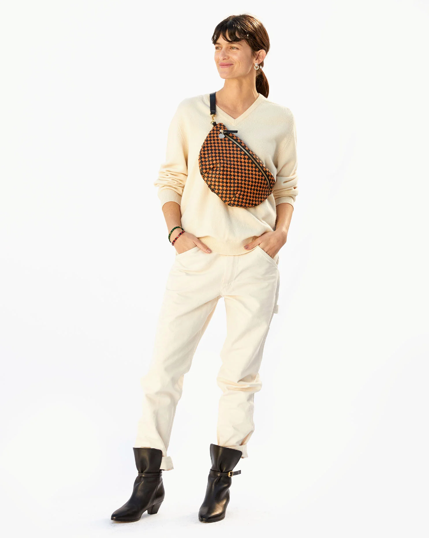 A woman stands confidently against a white background, embodying Arizona style in a cream sweater, white pants, black boots, and carrying a diagonally-slung Clare Vivier Grande Fanny Woven Checker handbag.