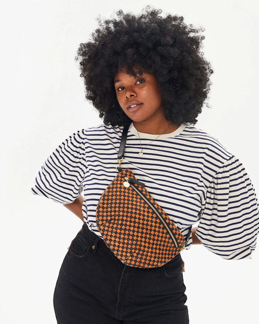 A woman with a voluminous curly hairstyle wearing a striped T-shirt and black jeans, holding a patterned Clare Vivier Grande Fanny Woven Checker, posing confidently against a white background in Arizona style.
