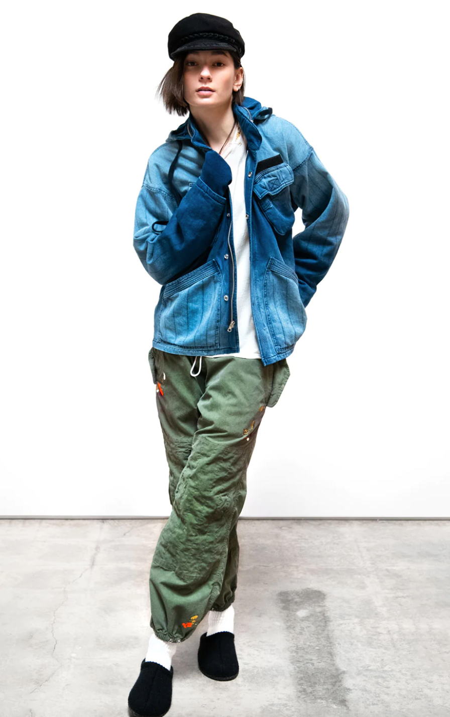 A young person sporting a stylish Free City (sparrow, LLC) ARTISTSWANTED INDIGO ZIP HOOD jacket - INDIGO2 over a white shirt and green embroidered pants stands confidently against a plain background, wearing a black cap and black socks.