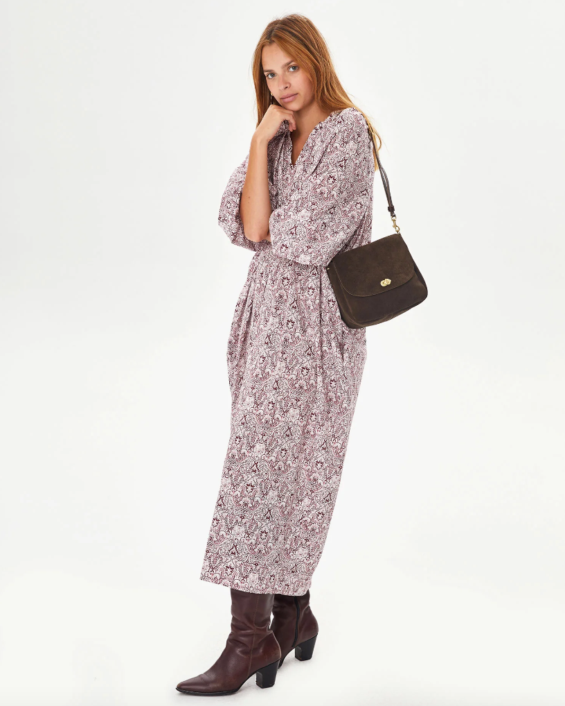 A woman stands against a white background, wearing a Clare Vivier Turnlock Louis midi dress, brown boots, and holding a shoulder bag, with her hand to her chin in a thoughtful pose.