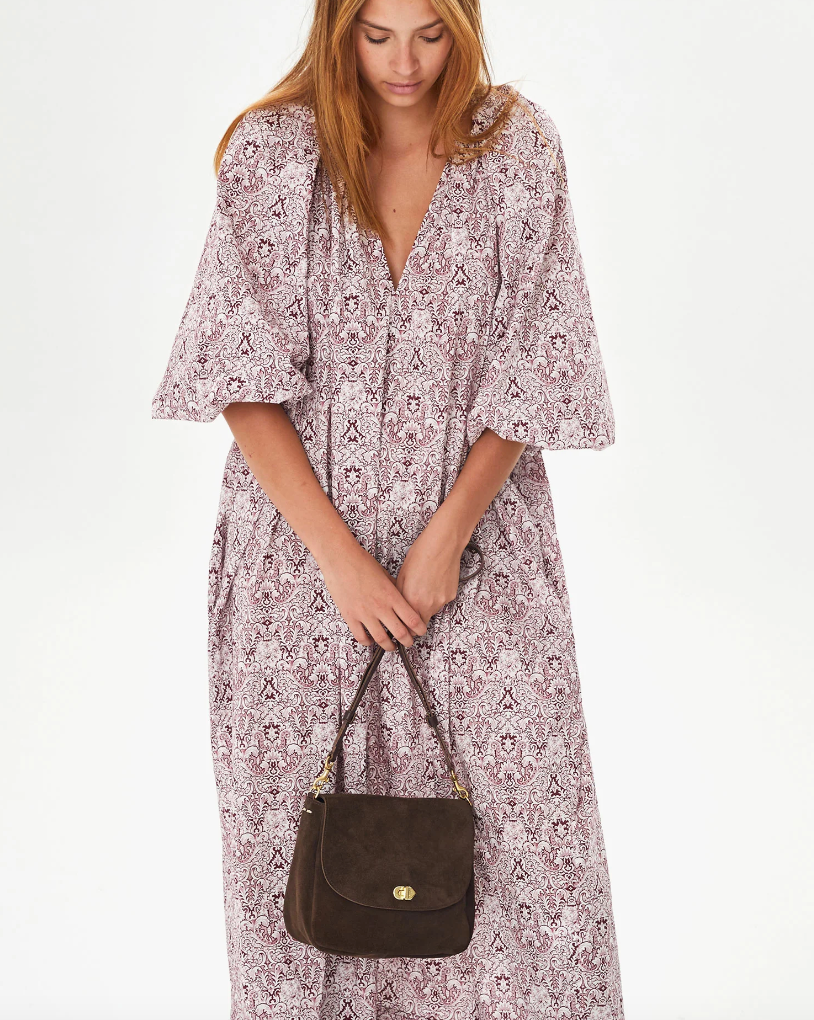 A woman in a flowy, floral print dress holds a dark brown Turnlock Louis shoulder bag by Clare Vivier. She faces down, highlighting the Arizona-style outfit and accessory.