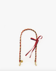 A multicolored Grosgrain in Chain friendship bracelet with red, yellow, and green threads, featuring a small golden charm and red ribbon tied in a bow, Clare Vivier style, isolated on a white background.