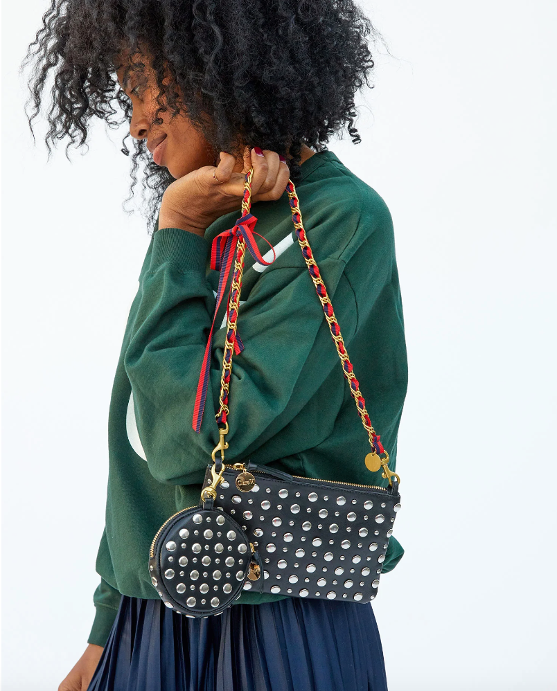 A woman with curly hair wearing a green sweater and a pleated skirt holds a studded bag with Clare Vivier&#39;s Grosgrain in Chain Shoulder Strap Navy/Red Grosgrain Chain over her shoulder, turned away from the camera, showcasing Arizona-inspired style.