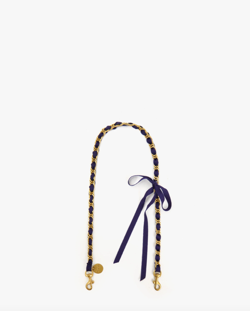 A stylish camera strap featuring the Grosgrain in Chain Shoulder Strap Navy/Red Grosgrain Chain by Clare Vivier, with a navy and yellow braided design with gold-tone hardware and a small blue bow tied near one end, displayed against a plain white Bungalow background.