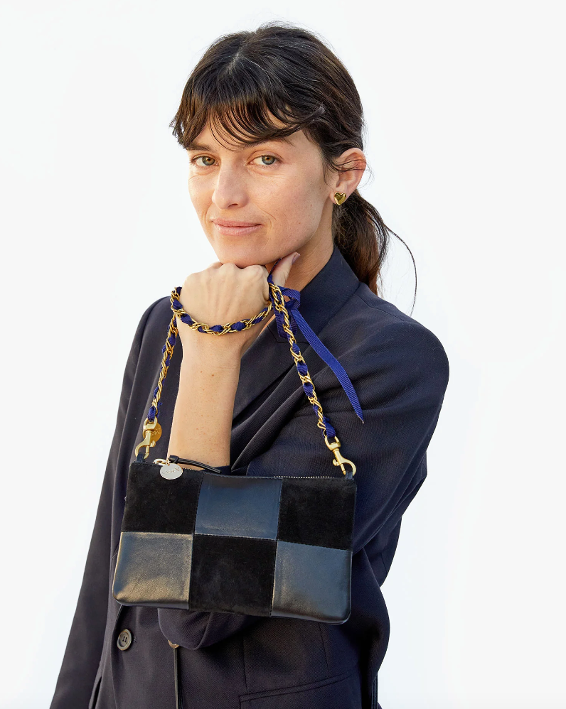 A woman with shoulder-length brown hair smiles subtly at the camera, holding a Clare Vivier Grosgrain in Chain Shoulder Strap Navy/Red Grosgrain Chain handbag on her forearm. She wears a dark blue Arizona-style outfit.
