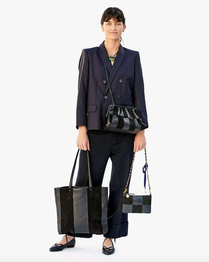 A woman in a dark blue business suit, carrying multiple bags, and wearing a green necklace, stands confidently against a plain Arizona background. She is also sporting the Grosgrain in Chain Shoulder Strap Navy/Red Grosgrain Chain from Clare Vivier.