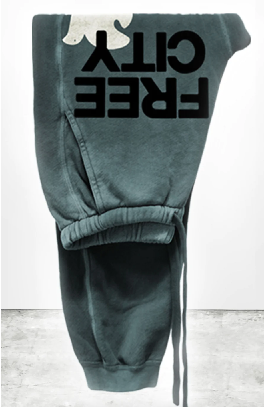 A teal sweatpant with the words "FREECITY" printed in black block letters, made from lightweight French terry, hanging on a wall. The image has a grayish concrete floor visible at the bottom.