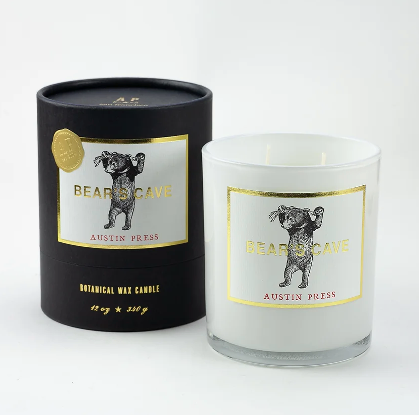 A Bear's Cave Candle by Austin Press, named "BEARSCAVE," displayed next to its cylindrical black packaging in Bungalow style. Both the candle and packaging feature a gold label with an illustration of a bear.